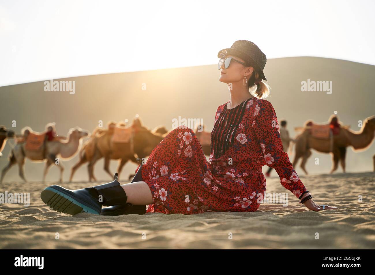 donna asiatica in abito rosso seduta nel deserto guardando la vista con caravan di cammelli e enorme duna di sabbia sullo sfondo Foto Stock