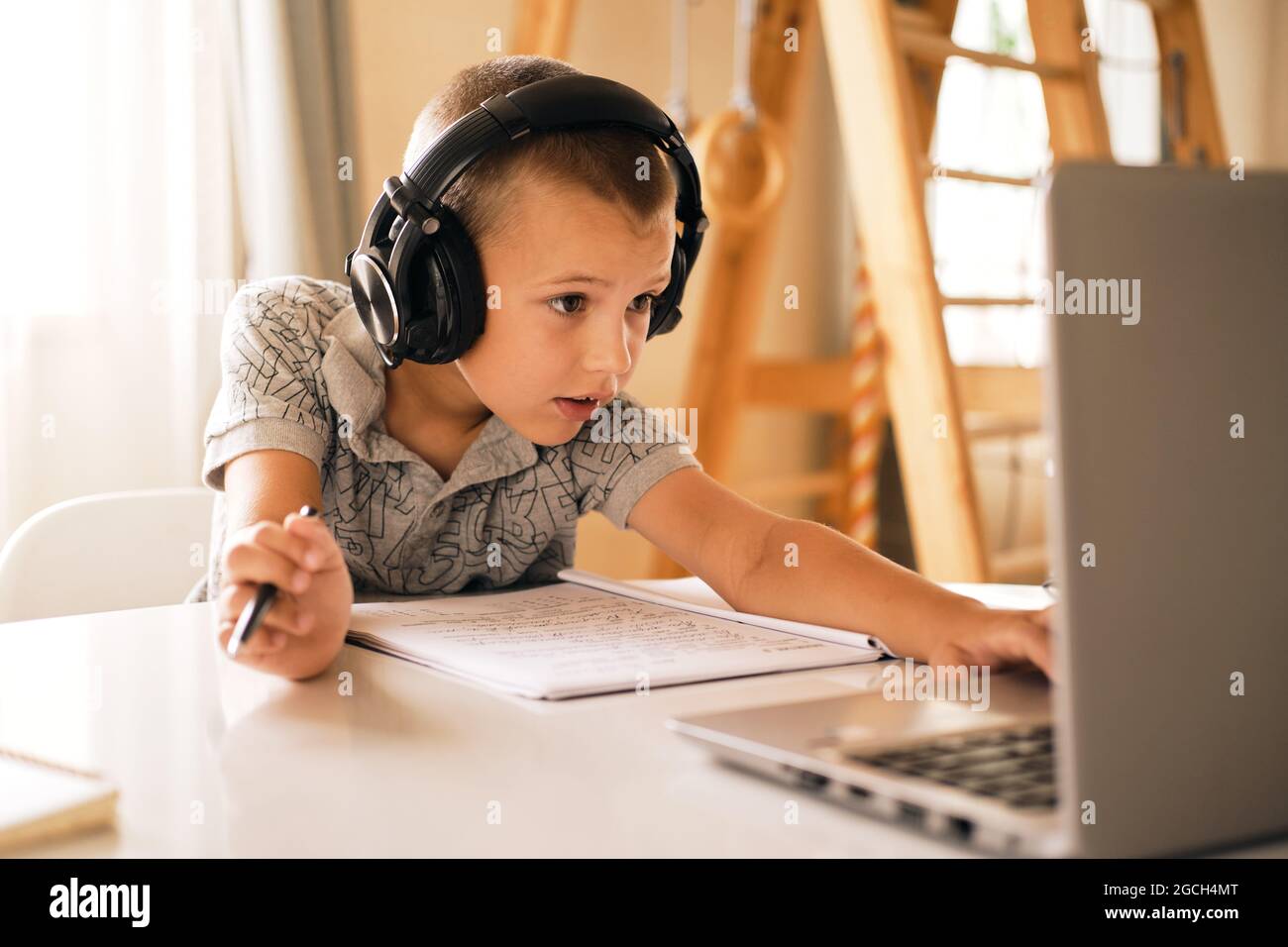 Ragazzo in cuffia che scrive nel notebook e usa il computer portatile mentre fa il lavoro scolastico a casa. Concetto di formazione online. Foto Stock