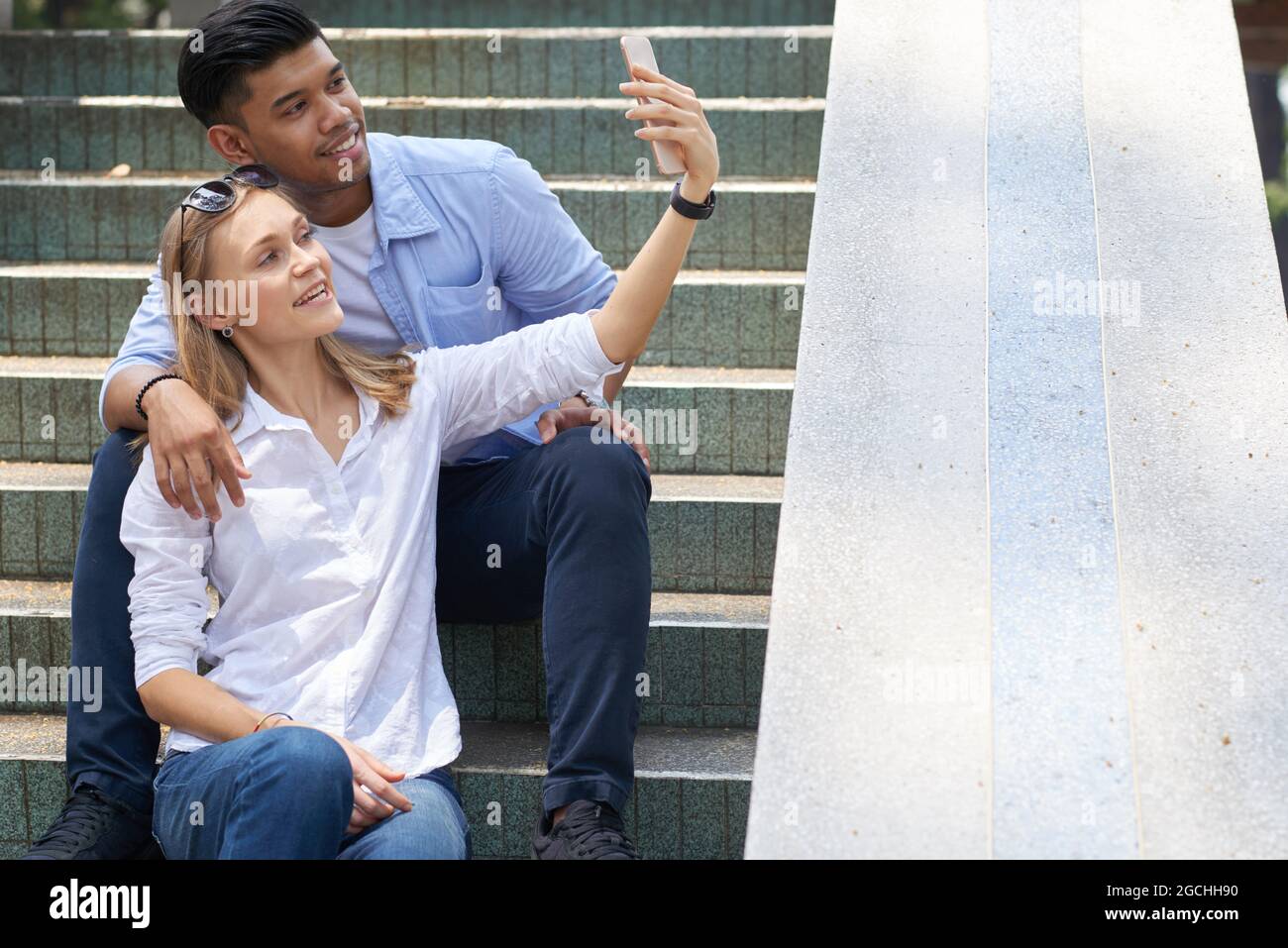 Felice giovane coppia varia seduta su passi e prendendo selfie per pubblicare sui social media Foto Stock