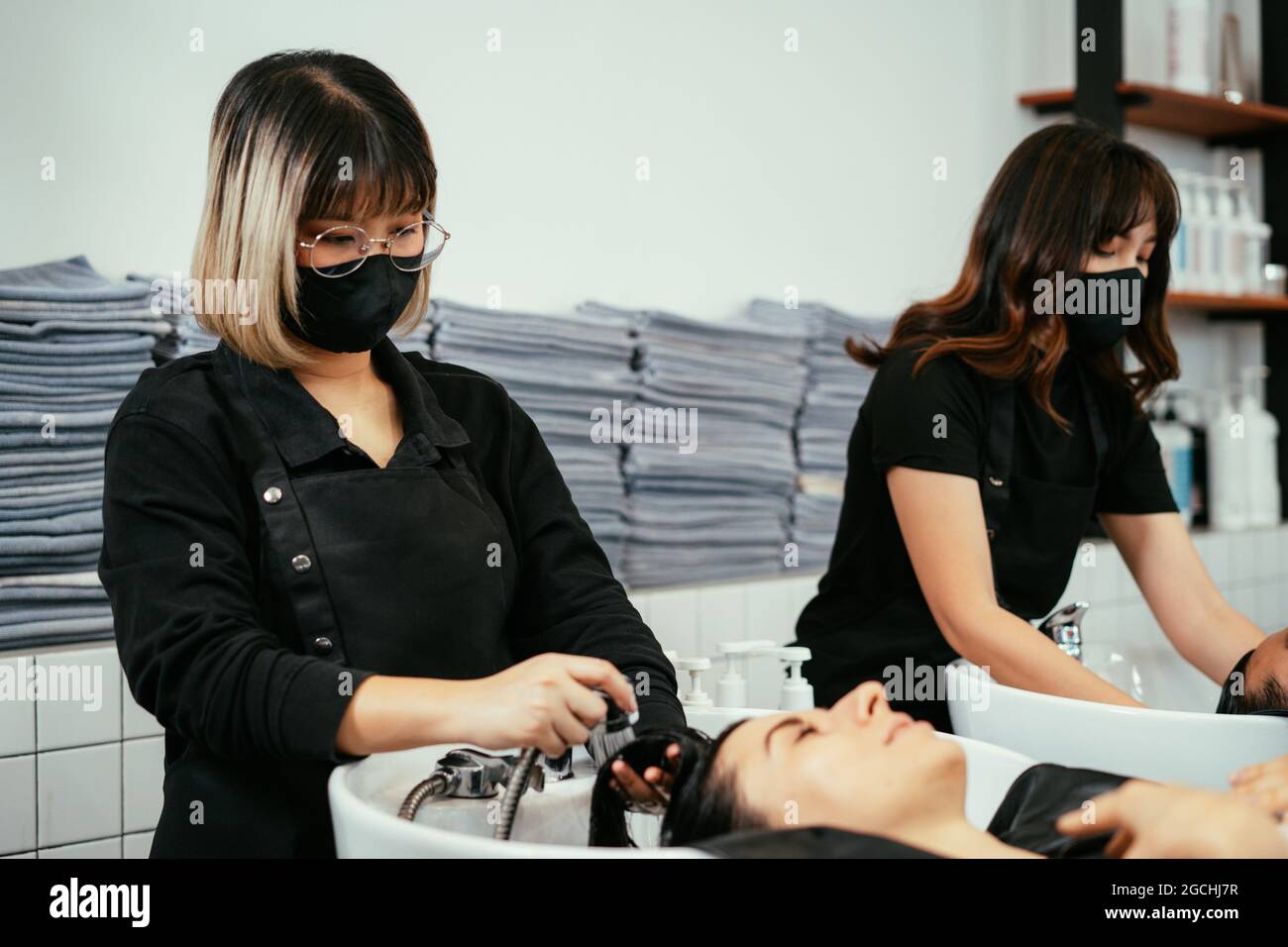 Giovane donna asiatica parrucchiere indossare covid-19 maschera protettiva viso lavaggio e risciacquo capelli bagnati di giovane donna cliente in salone moderno Foto Stock