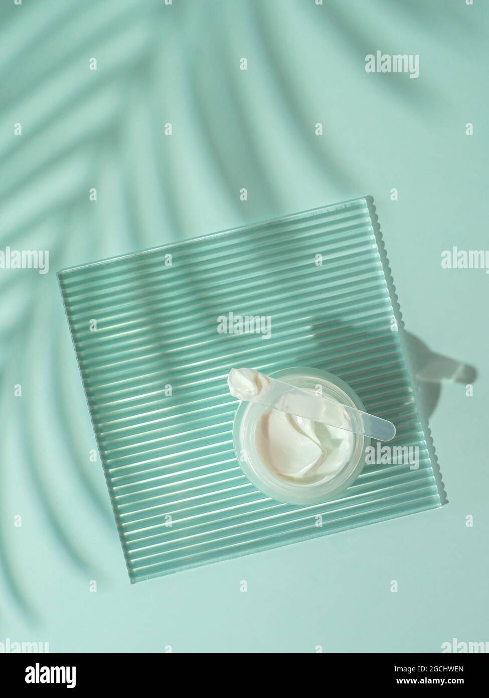 Moustirizer crema cosmetica, lastra acrilica a coste trasparenti, ombre di foglie di palma tropicali su sfondo blu. Aprire il vaso rotondo di vetro, crema di swirls estetica, spatola. Vista dall'alto piatta. Spazio di copia. Verticale Foto Stock