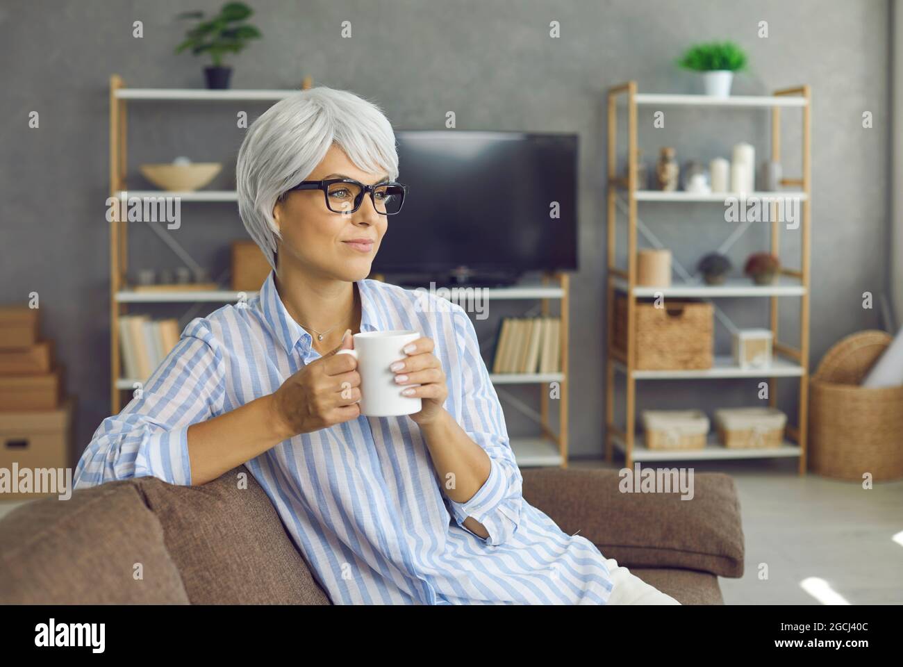 Signora matura felice rilassarsi sul divano, gustando una buona tazza di tè, guardando via e pensando Foto Stock