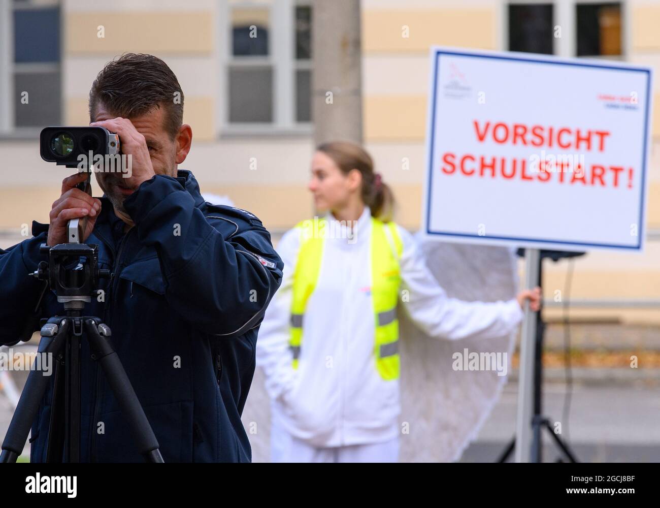 09 agosto 2021, Brandeburgo, Potsdam: Il sovrintendente capo della polizia Radtke controlla la velocità dei veicoli su Yorckstraße, che è designato per 30 km/h, alla scuola primaria Max Dortu utilizzando la misurazione laser. Sullo sfondo c'è una giovane donna in costume da angelo con un cartello che dice 'attenzione, inizio della scuola'. Nei primi quaranta giorni dopo l'inizio della scuola dopo le vacanze estive, la polizia intende intensificare i controlli del traffico stradale in tempi prioritari. Allo stesso tempo, la campagna di sicurezza stradale 'Lieber sicher. Lieber leben.' è stato lanciato per promuovere la considerazione tra ciclisti e autisti Foto Stock