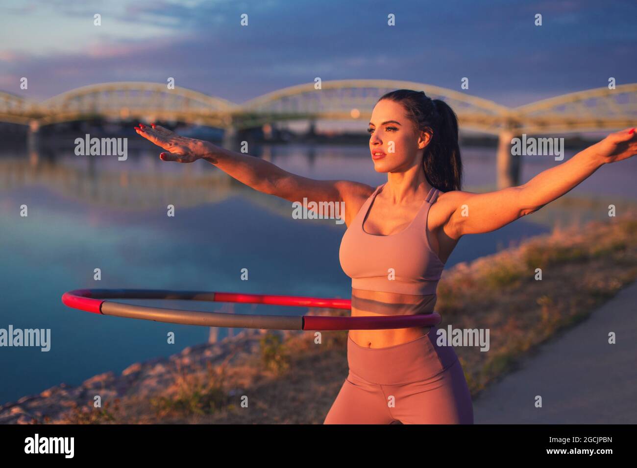Giovane donna che fa l'esercitazione di hula hoop al lungofiume nel tramonto Foto Stock