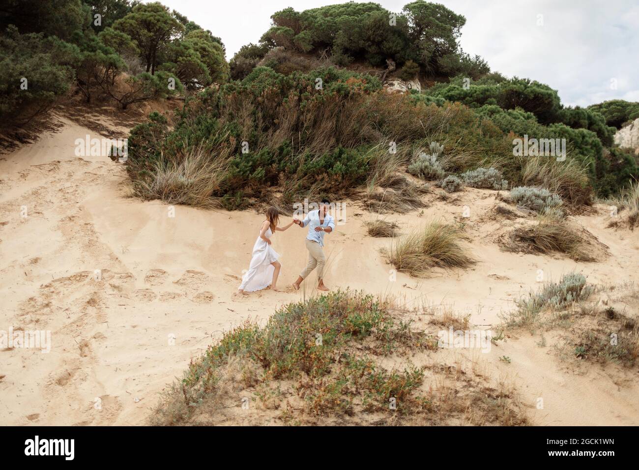 Felice ponte multirazziale e sposo tenendo le mani e correndo lungo la collina di sabbia il giorno di nozze in natura Foto Stock
