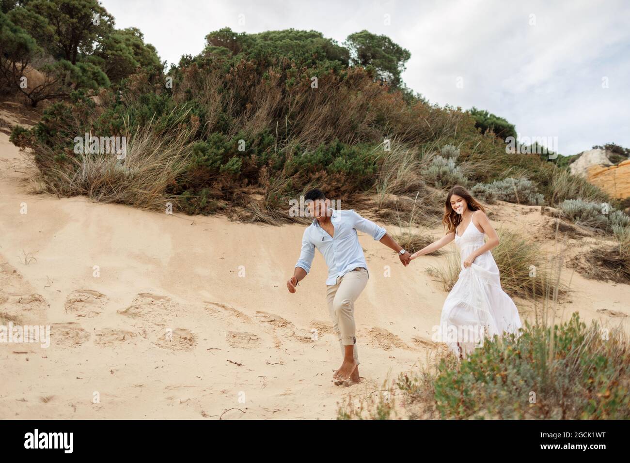 Felice ponte multirazziale e sposo tenendo le mani e correndo lungo la collina di sabbia il giorno di nozze in natura Foto Stock