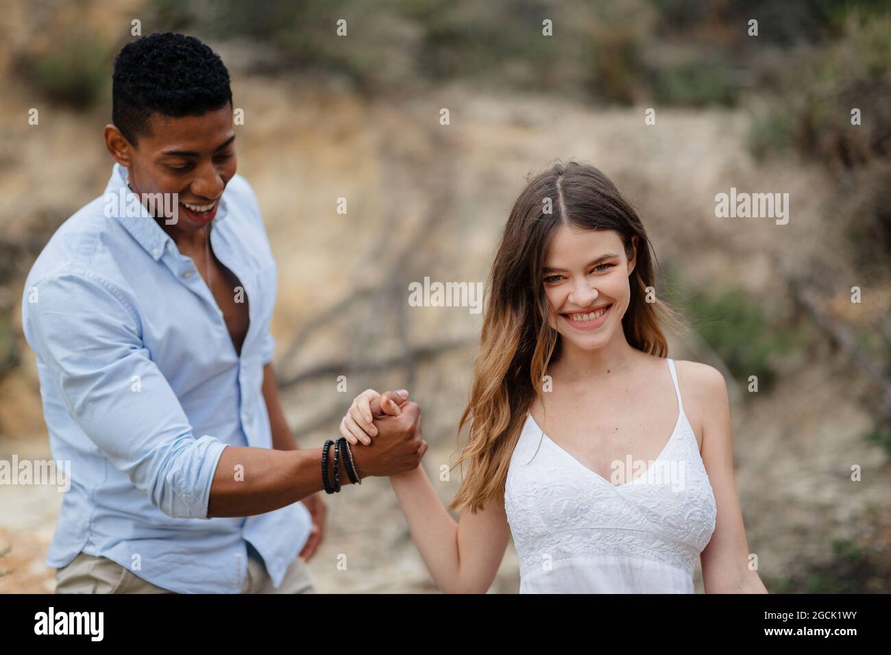 Felice ponte multirazziale e sposo tenendo le mani e camminando lungo la collina di sabbia il giorno di nozze in natura Foto Stock