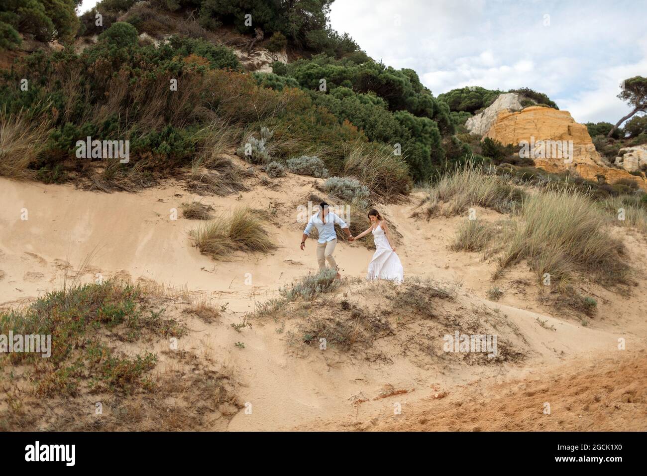 Felice ponte multirazziale e sposo tenendo le mani e correndo lungo la collina di sabbia il giorno di nozze in natura Foto Stock
