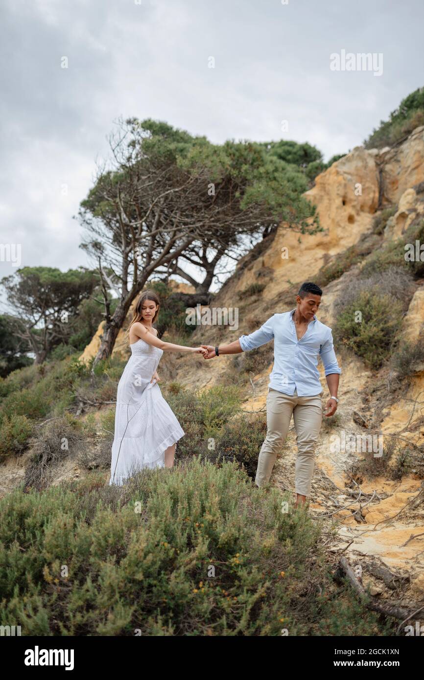 Felice ponte multirazziale e sposo tenendo le mani e camminando lungo la collina di sabbia il giorno di nozze in natura Foto Stock