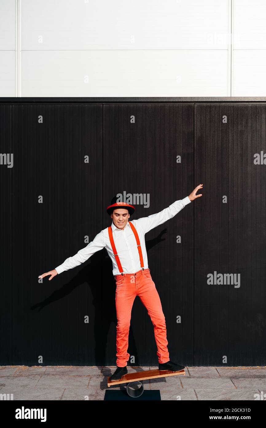 Corpo pieno di artista maschile circo in pantaloni rossi e cappello che esegue trucco sulla tavola di bilanciamento contro parete nera Foto Stock