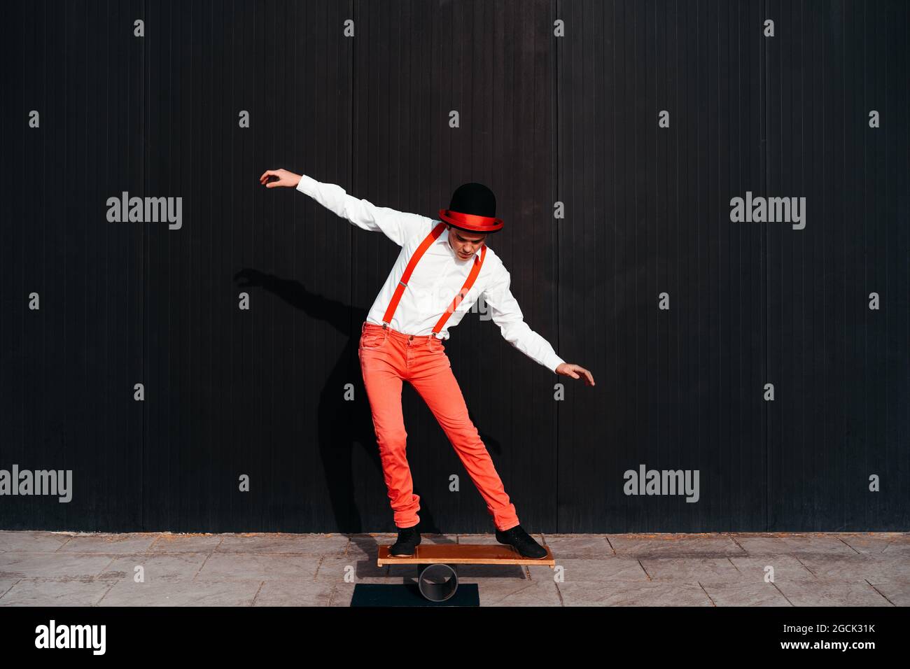 Corpo pieno di artista maschile circo in pantaloni rossi e cappello che esegue trucco sulla tavola di bilanciamento contro parete nera Foto Stock