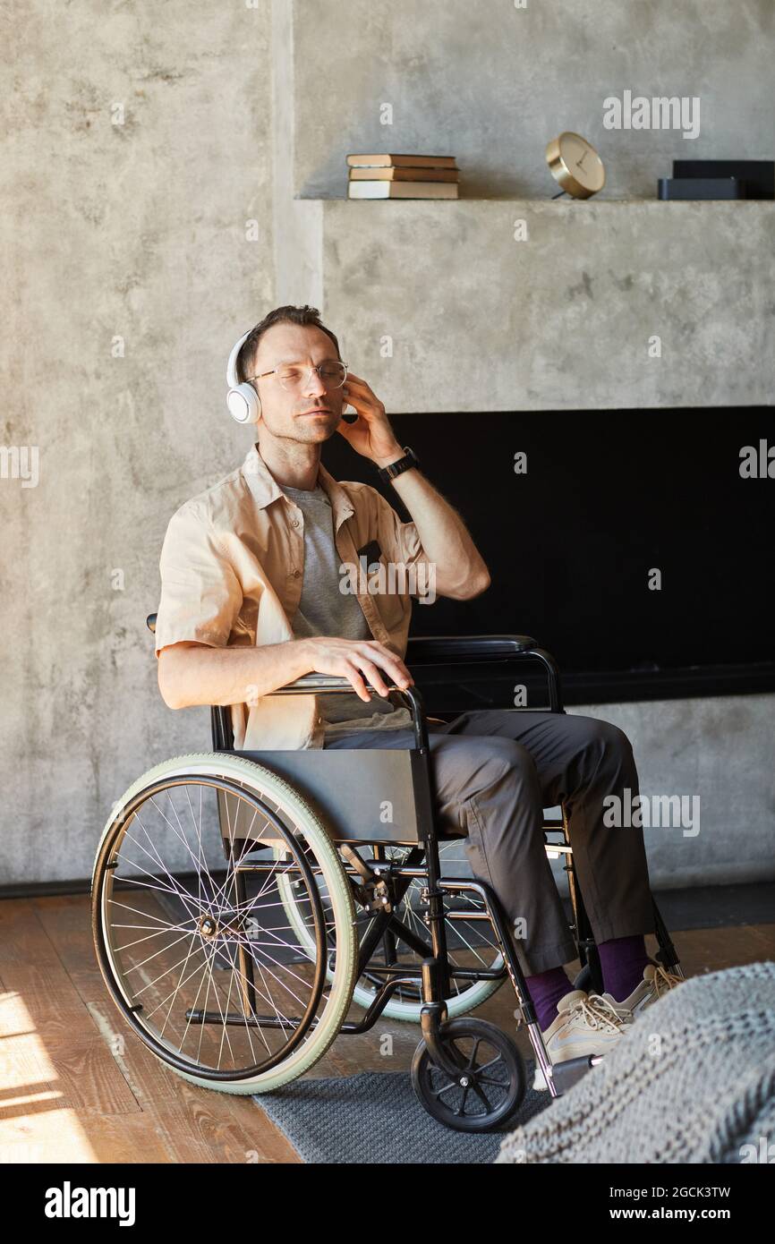 Ritratto verticale a lunghezza intera dell'uomo adulto in sedia a rotelle che ascolta la musica a casa Foto Stock