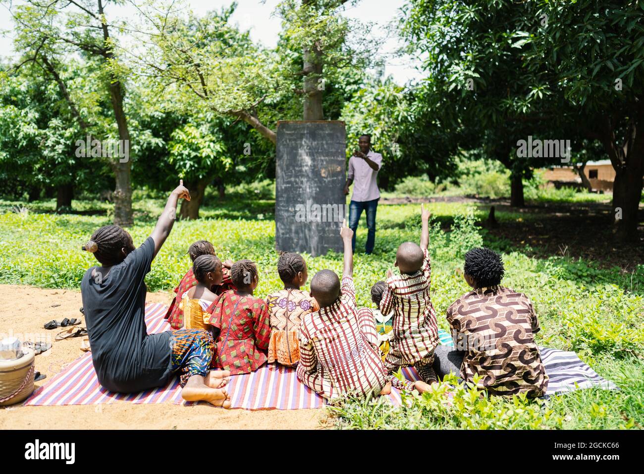 Gruppo misto di bambini neri che si siedono all'aperto, con ansia di volontariato per rispondere alle domande del loro insegnante durante la lezione in una scuola di comunità africana Foto Stock