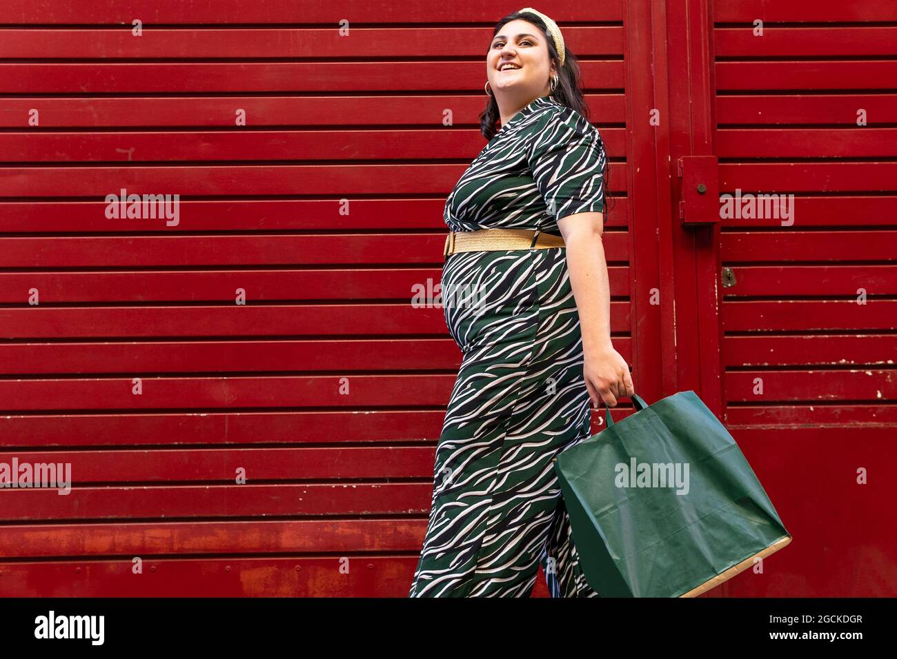 Vista laterale ad angolo basso della giovane donna giovane e curvy in un abito elegante con stampa geometrica che trasporta borse per la spesa mentre si trova vicino al muro a strisce rosse in c. Foto Stock