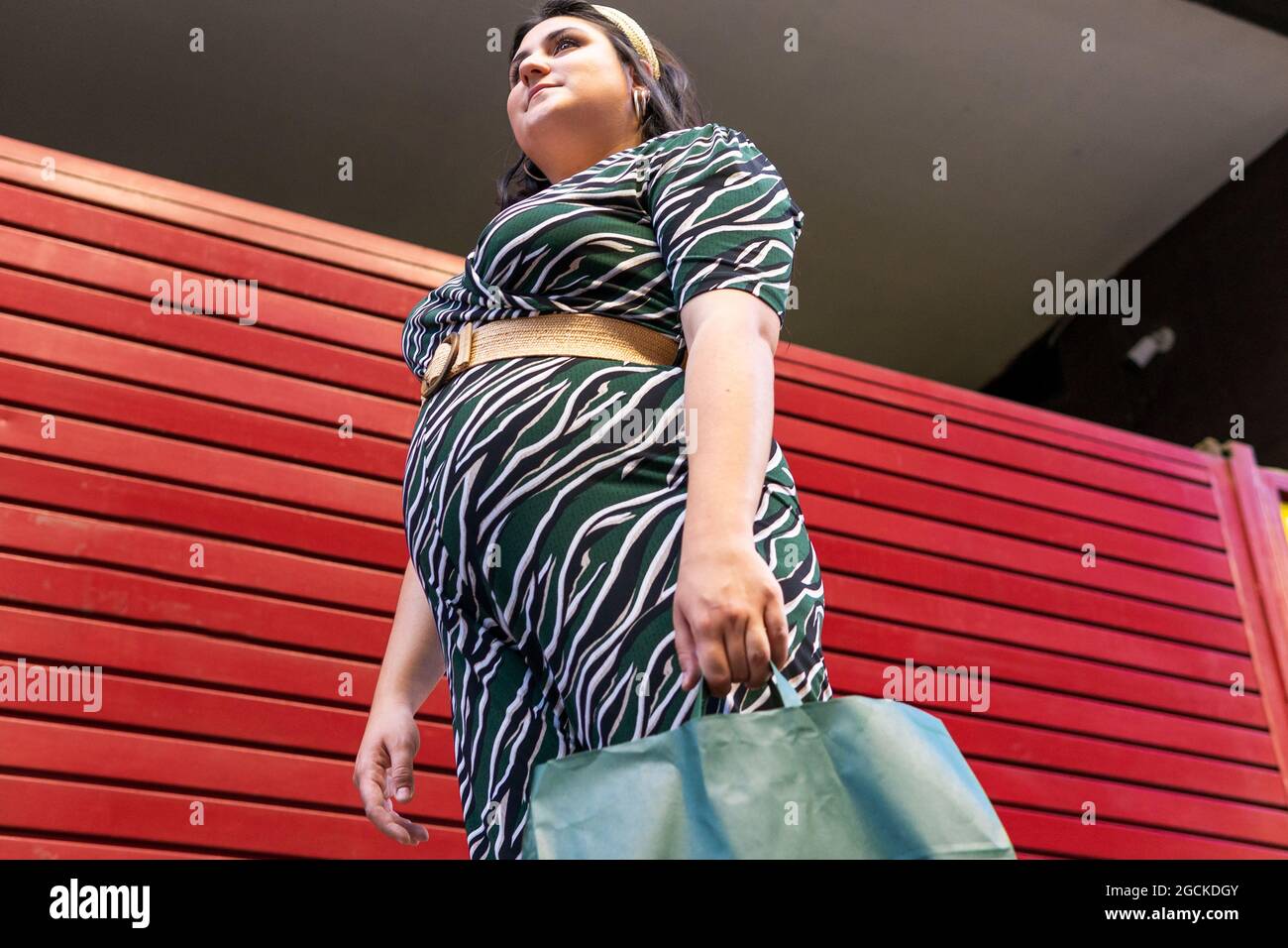 Vista laterale ad angolo basso della giovane donna giovane e curvy in un abito elegante con stampa geometrica che trasporta borse per la spesa mentre si trova vicino al muro a strisce rosse in c. Foto Stock