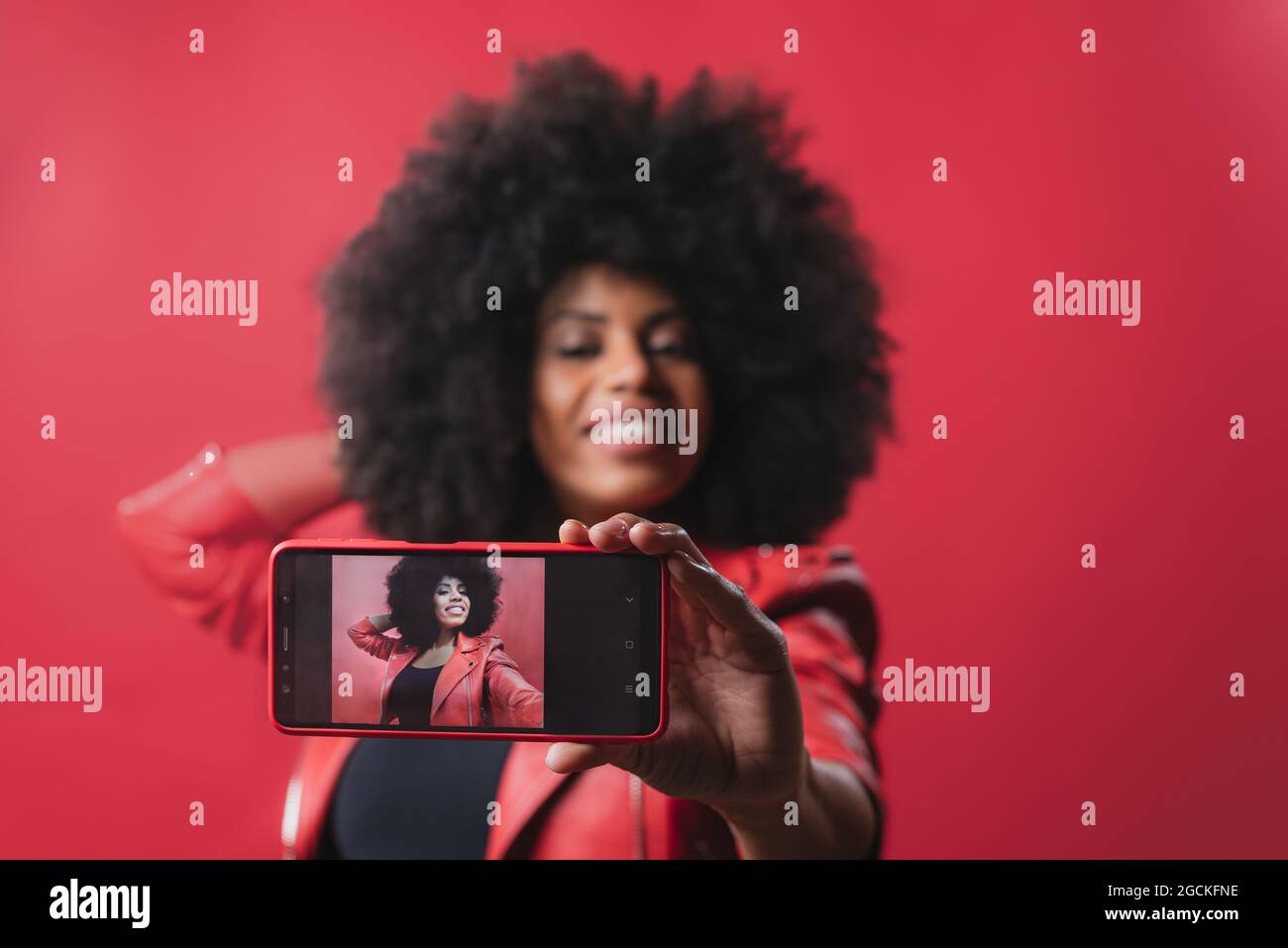 Femmina afro-americana sorridente con acconciatura Afro che si autoritratto sul telefono cellulare su sfondo rosso in studio Foto Stock