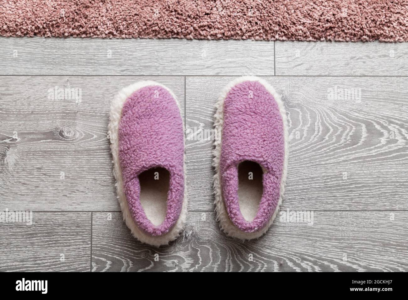 Le accoglienti pantofole in lana si trovano su un pavimento in legno grigio di fronte a un tappeto rosa Foto Stock