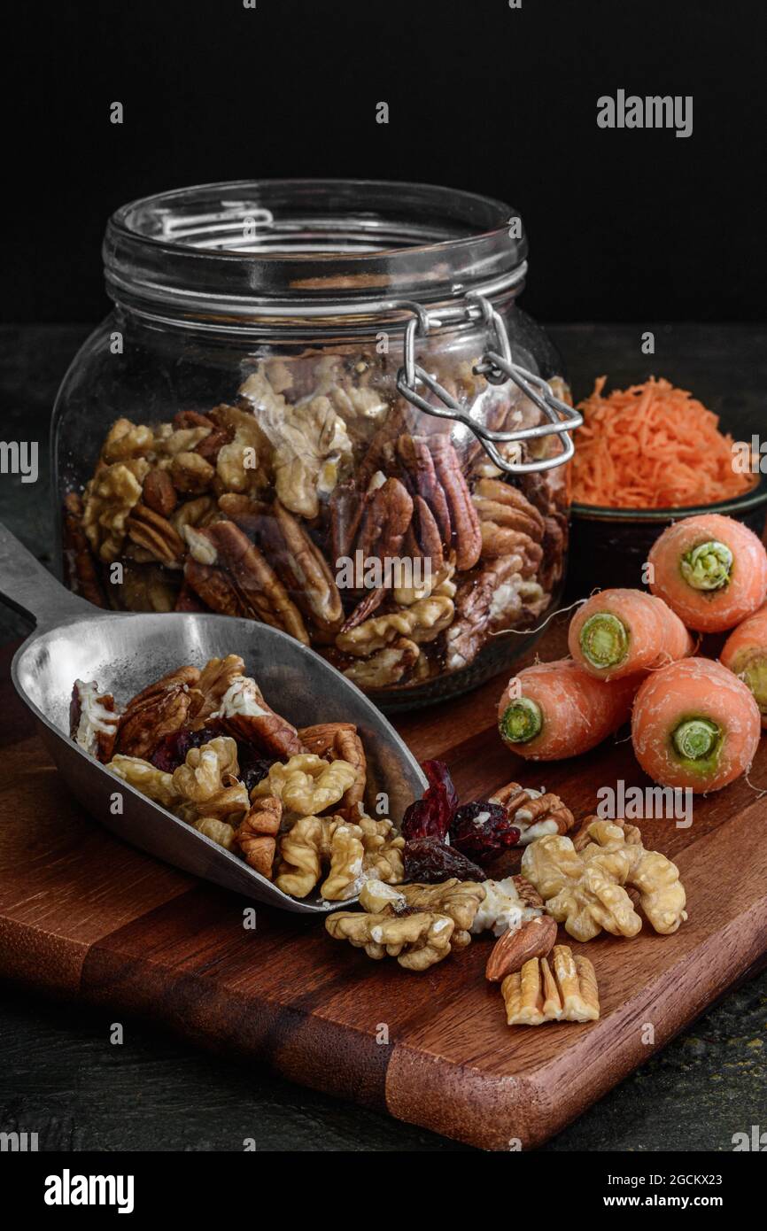 Vasetto di vetro con noci e frutta secca su sfondo rustico scuro accanto alle carote, ingredienti per preparare una torta di carote Foto Stock