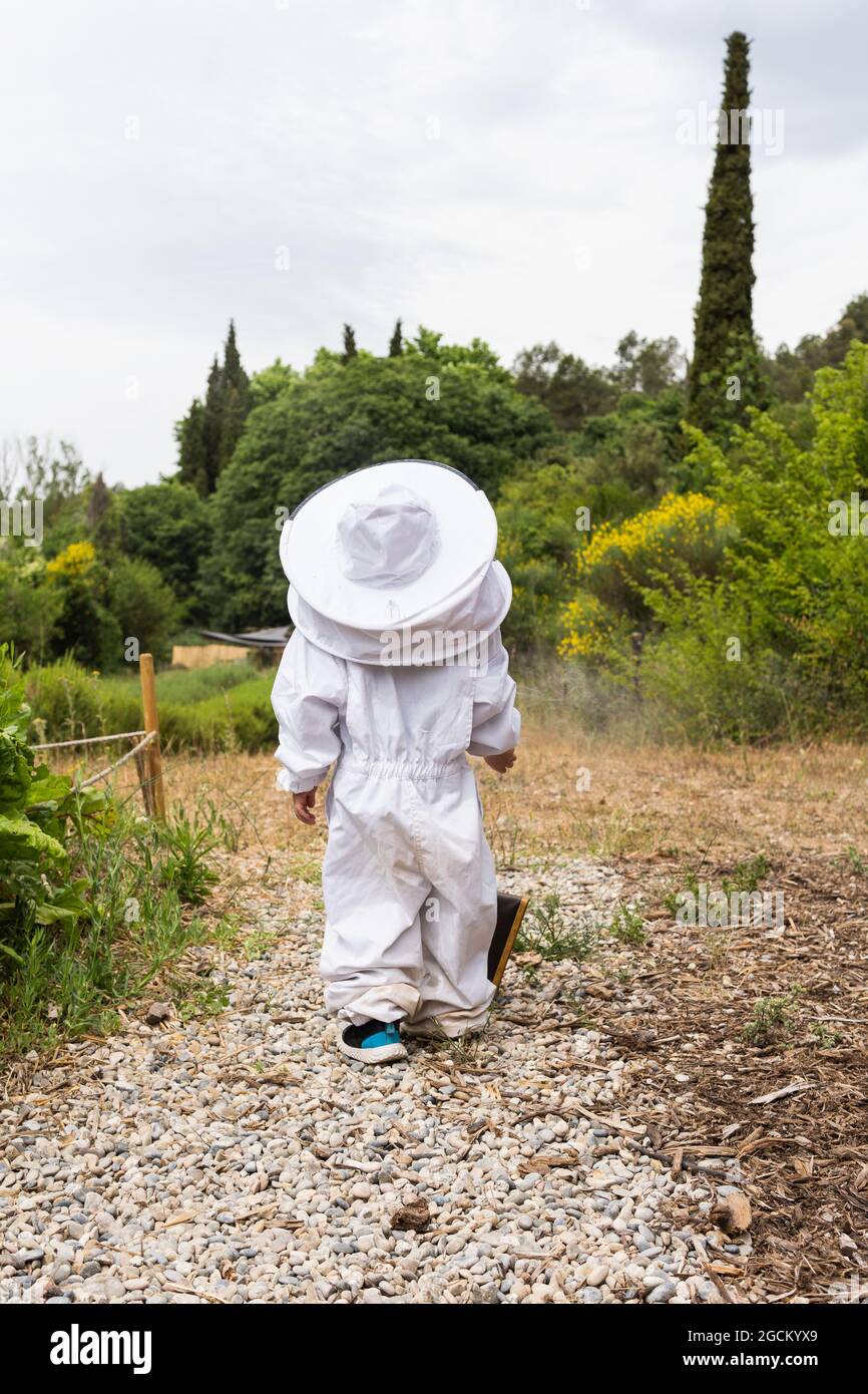 Vista posteriore di bambino irriconoscibile in costume protettivo bianco apicoltore in piedi vicino al fumatore di api in metallo nel villaggio Foto Stock