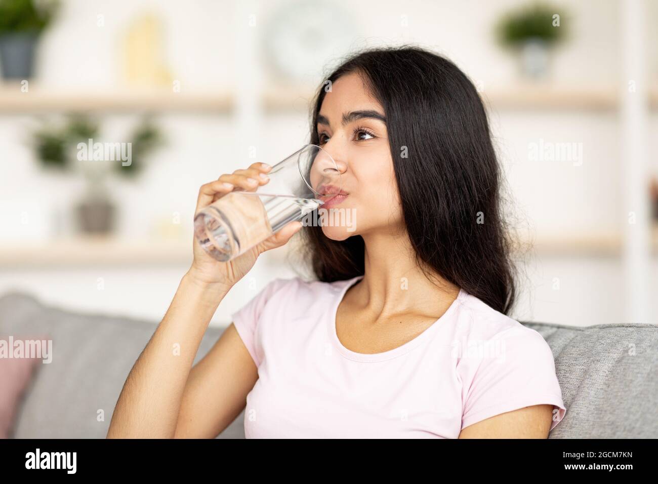 Concetto di idratazione. Attraente giovane donna indiana che beve acqua dal vetro sul divano in soggiorno Foto Stock