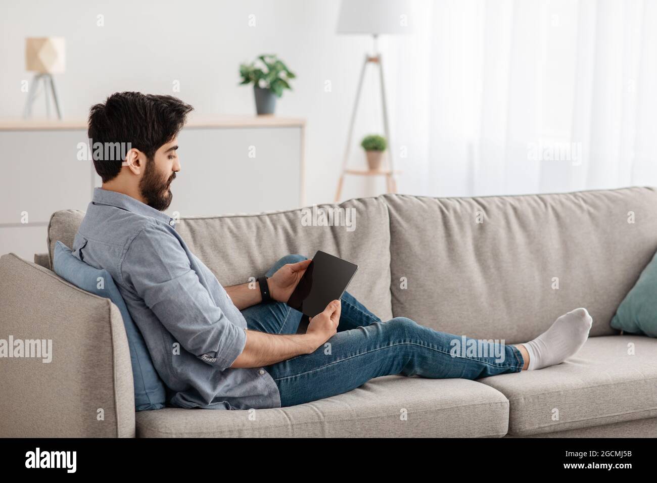 Giovane arabo che riposa con un tablet digitale, che si distende sul divano in soggiorno, spazio per la copia Foto Stock