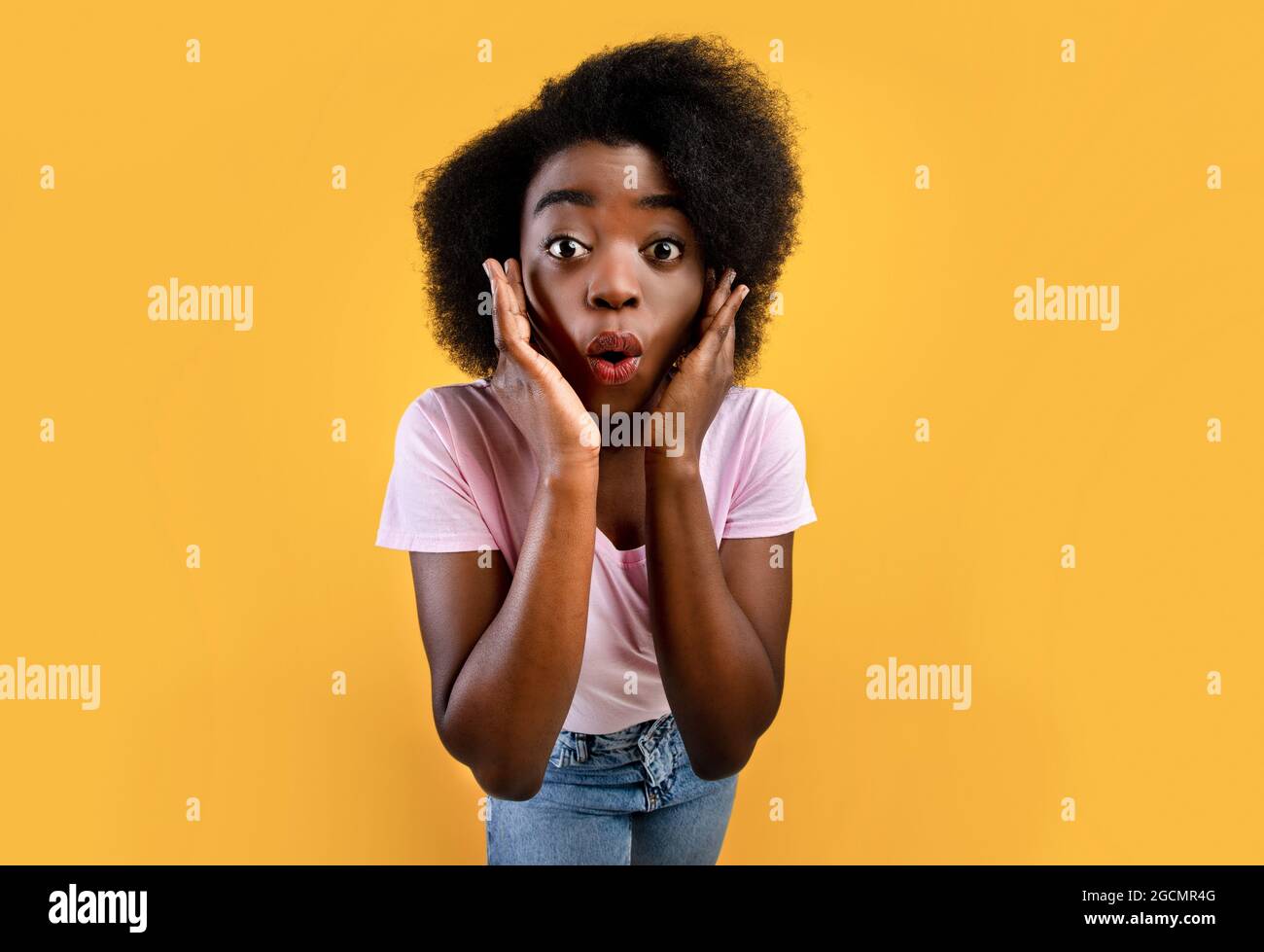 Incredibile. Scioccata donna afroamericana con bocca aperta e mani su guance che posano su sfondo giallo Foto Stock