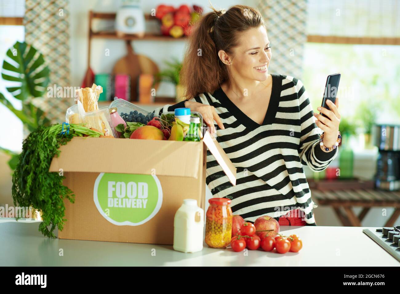 Consegna di cibo. Donna giovane felice con scatola di cibo in cucina utilizzando applicazioni smartphone. Foto Stock