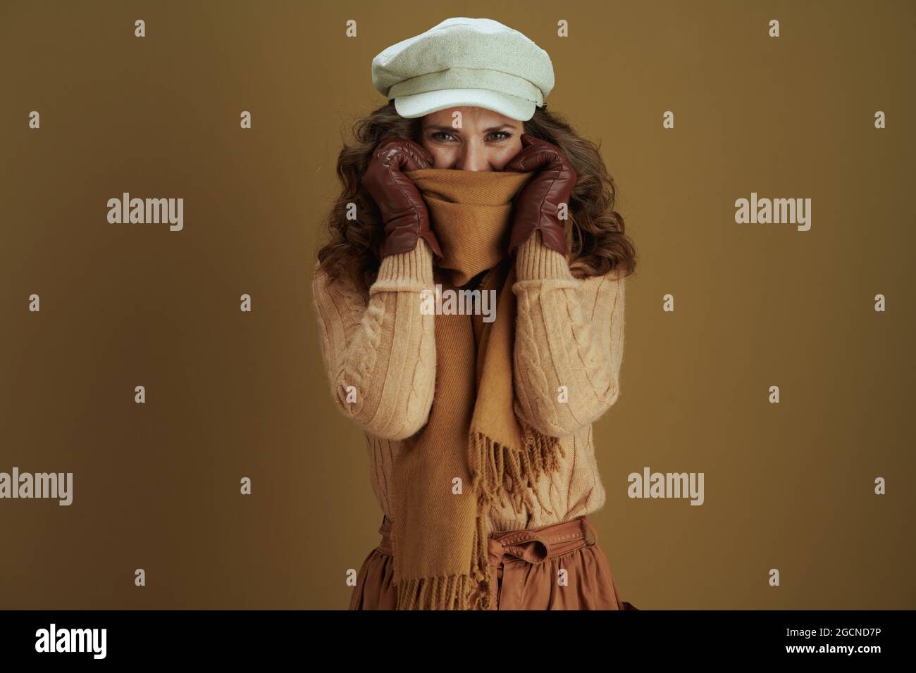 Ciao novembre. Donna moderna sorridente in pullover con guanti in pelle avvolgente in una sciarpa su sfondo beige. Foto Stock