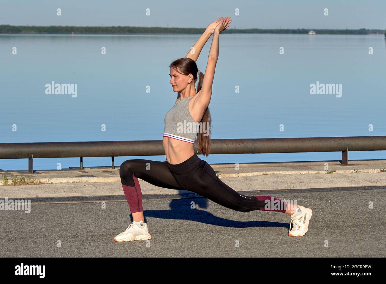 Una giovane donna fa un riscaldamento sul molo la mattina presto, sullo sfondo del fiume Foto Stock