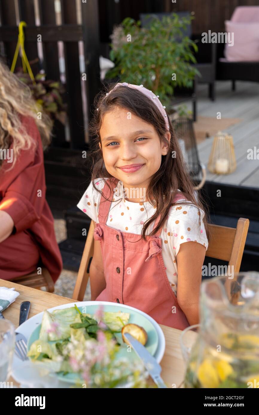 Adorabile ragazza sorridente mentre si siede al tavolo servito durante la cena all'aperto Foto Stock