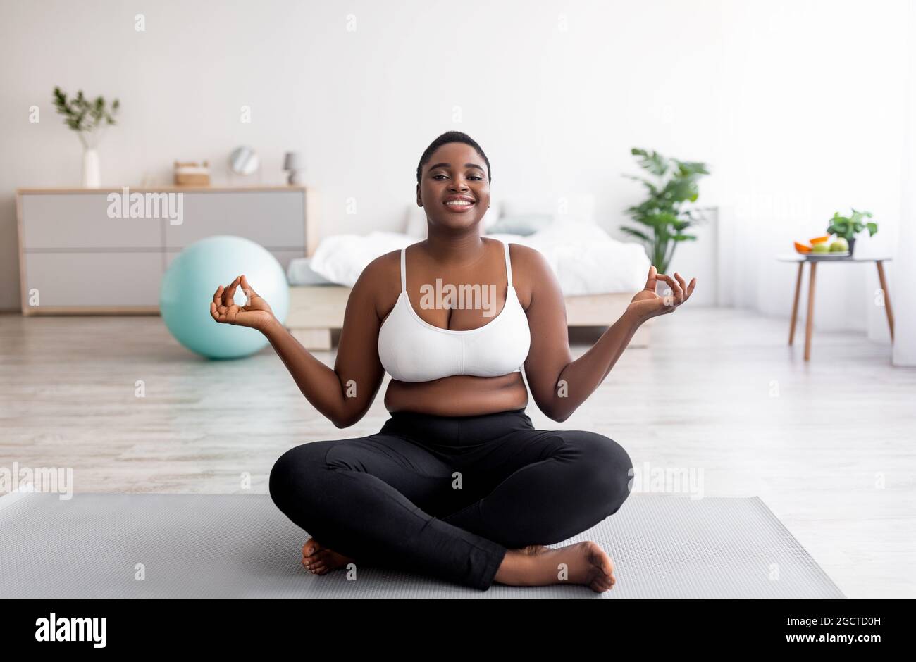 Donna nera sovrappeso seduta sul tappeto sportivo in posa lotus, meditando, facendo pratica yoga a casa Foto Stock