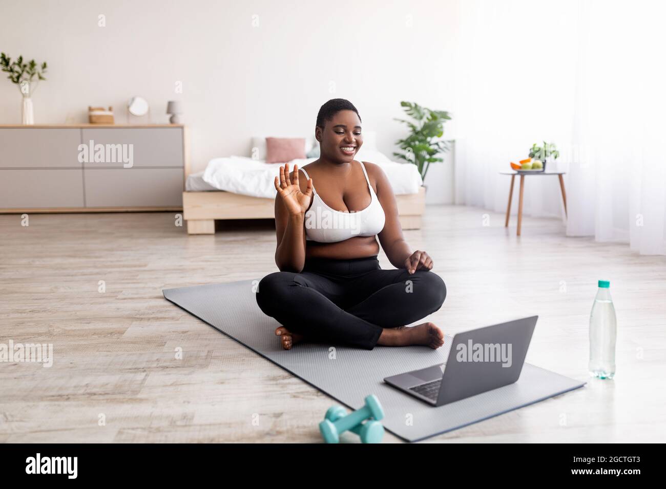 Donna nera in sovrappeso seduta sul tappetino yoga di fronte al computer portatile, saluto personal trainer, agitando alla webcam, spazio per la copia Foto Stock
