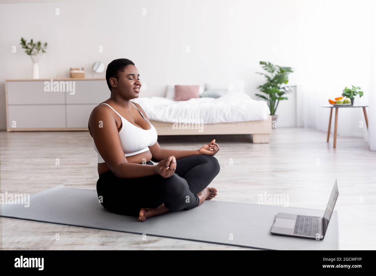 Pump donna nera che ha meditazione online o lezione di yoga su computer portatile, seduta con gli occhi chiusi in lotus posa a casa Foto Stock