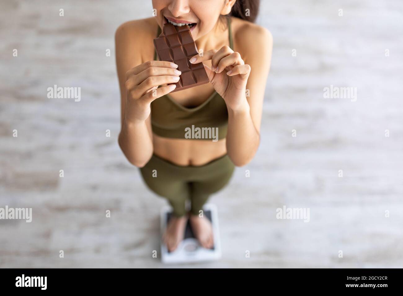 Fallimento di dieta, concetto di pasto di frode. Sopra la vista della donna indiana che mangia cioccolato mentre si sta in piedi su una bilancia Foto Stock