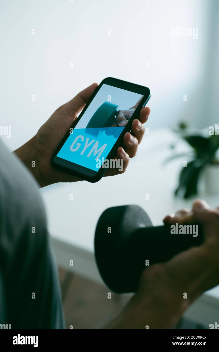 un giovane caucasico solleva un manubri nel soggiorno di casa mentre guarda al suo smartphone, che legge la palestra nel suo schermo Foto Stock
