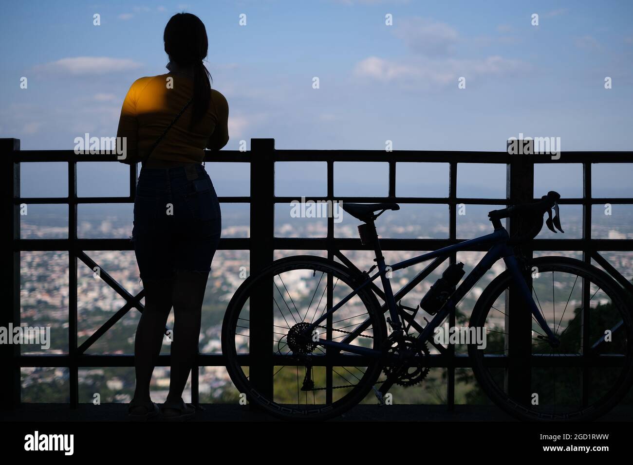 Giovane donna sportiva con la sua bicicletta è in piedi su un punto panoramico in collina di fronte al bellissimo scenario di una città lontana (Chiang mai) Foto Stock