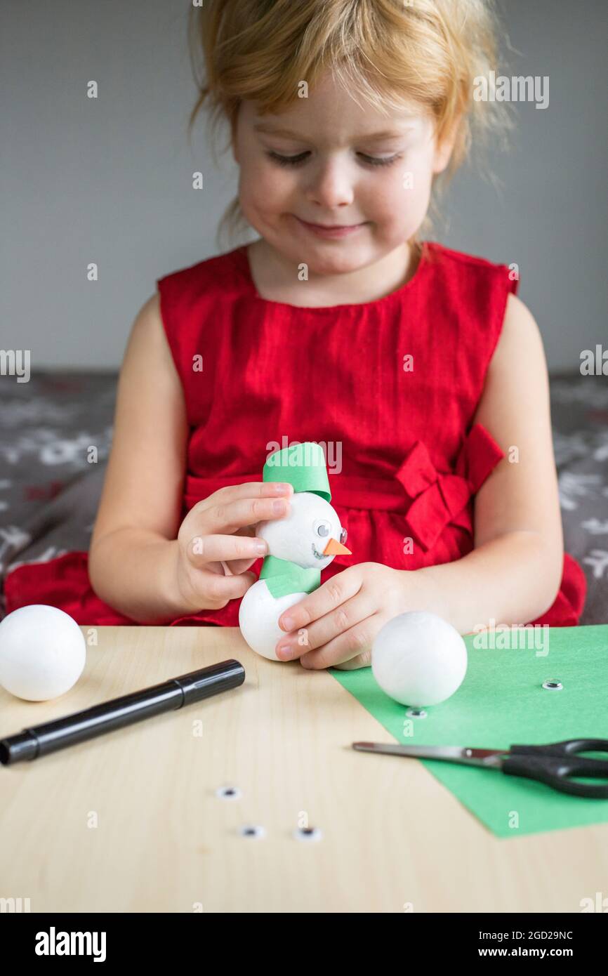 Natale bambini artigianato attività a casa. Piccola ragazza sorridente in abito rosso che fa pupazzo di neve. Il bambino è occupato a giocare. Concetto DIY. Foto Stock