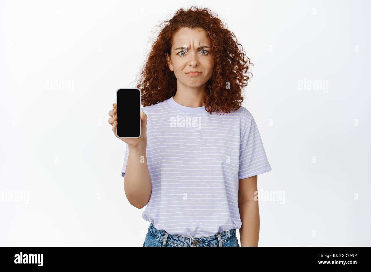 Immagine di giovane donna che mostra lo schermo del telefono cellulare con scettica, deluso espressione del volto, lamentando l'applicazione, sfondo bianco Foto Stock