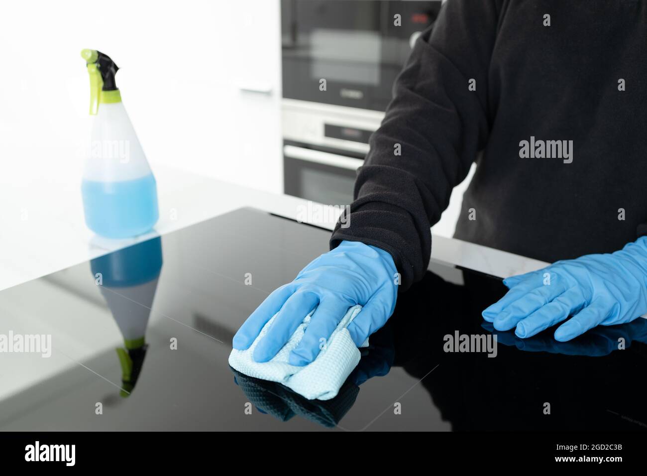 Mani in guanti protettivi pulizia di una stufa elettrica moderna con straccio di una cucina minimalista Foto Stock