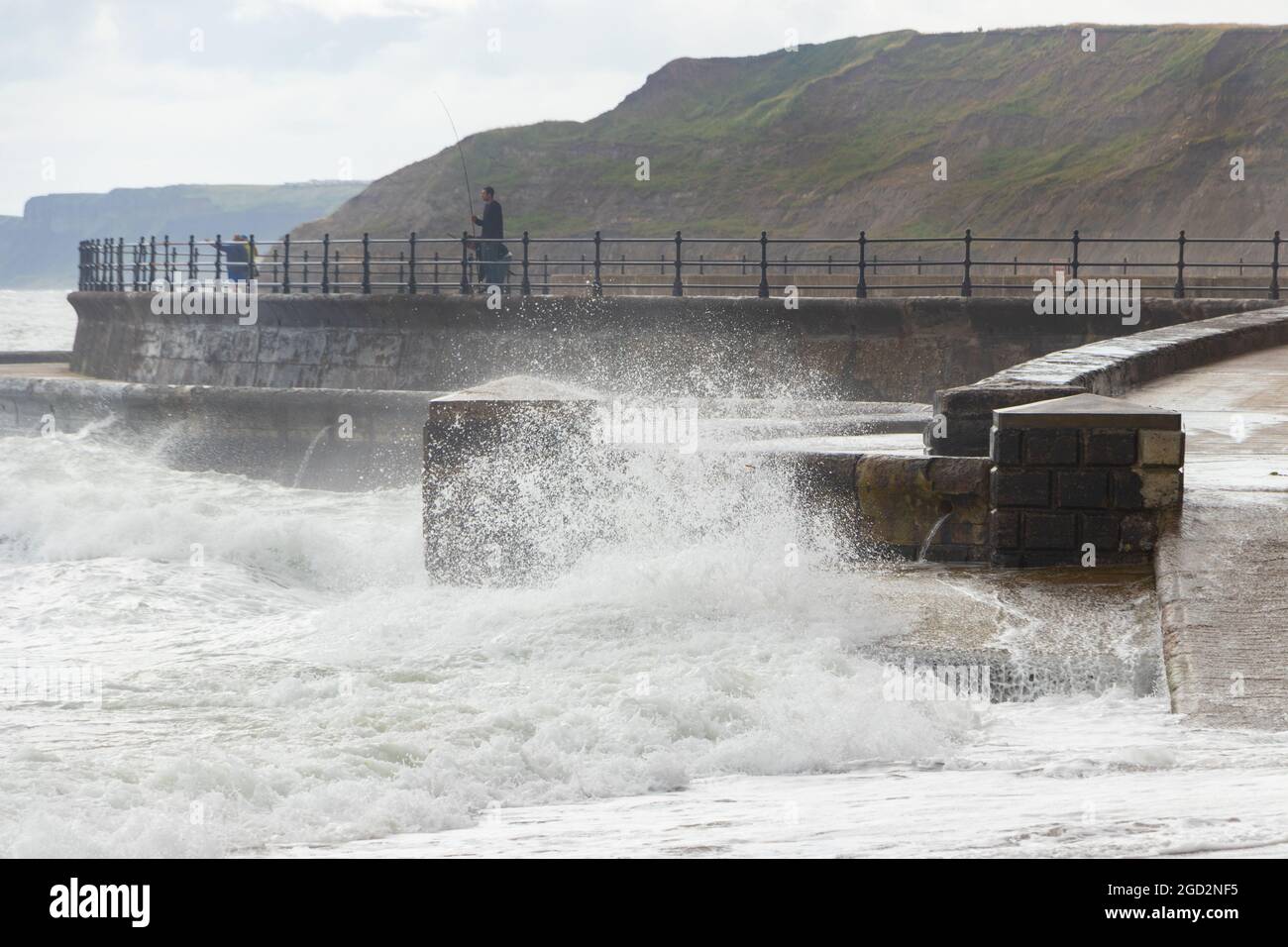 Uomo pesca con onde che si infrangono contro le difese marine nello Scarborough Yorkshire Foto Stock