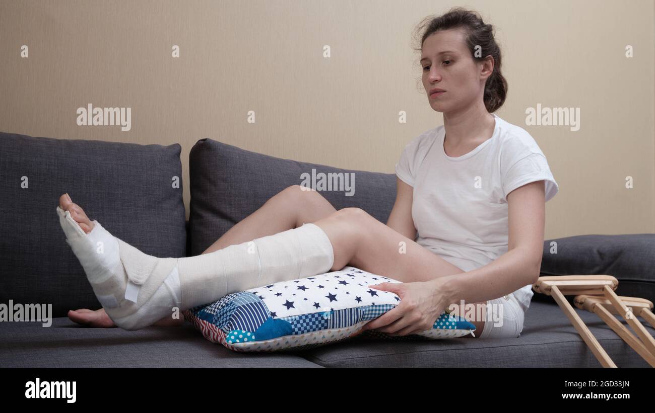 donna con un calco di gesso si siede su un divano, un cuscino è posto sotto una gamba rotta. Foto Stock