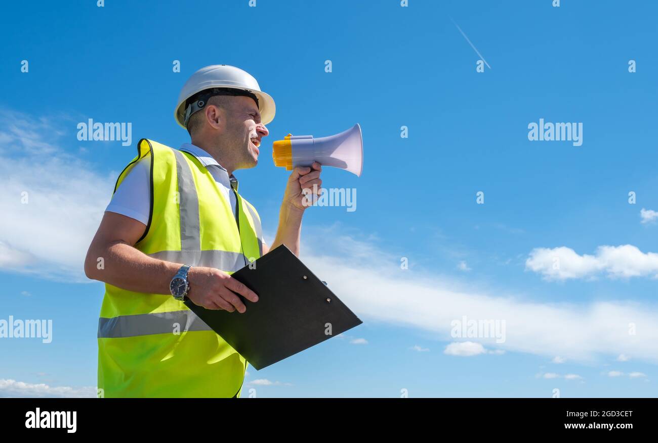 Il costruttore in un casco bianco urla in un megafono contro uno sfondo blu del cielo Foto Stock
