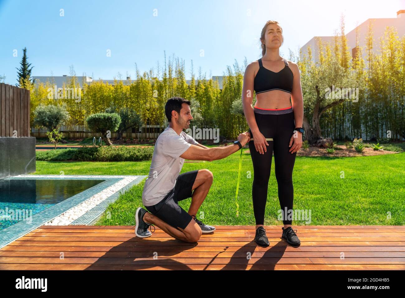Un uomo europeo, personal trainer, misura la gamba del cliente, l'anca per il progresso nella formazione, per la perdita di peso. In estate, in giardino con piscina. Foto Stock