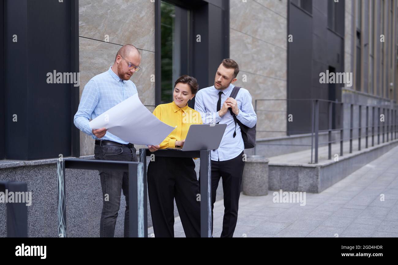 I progettisti o il team di architetti che guardano il progetto e il notebook come presentazioni all'aperto. Uomini d'affari, uomini e donne in abiti casual in piedi fuori, progetto di brainstorming Foto Stock