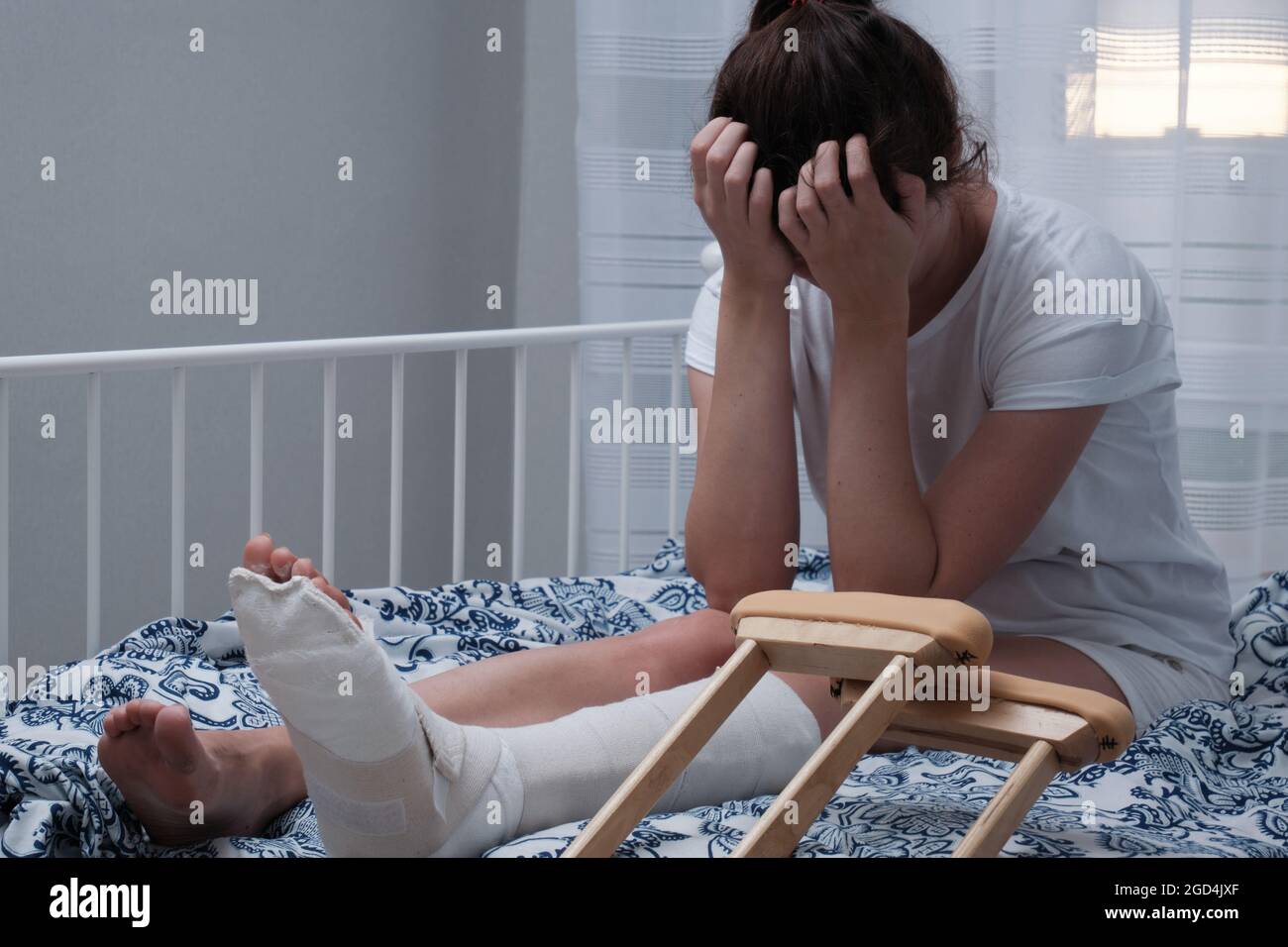 Una donna con un getto sulla gamba si siede sul letto, grida e copre il viso con le mani. Riabilitazione dopo trauma fisico. Foto Stock