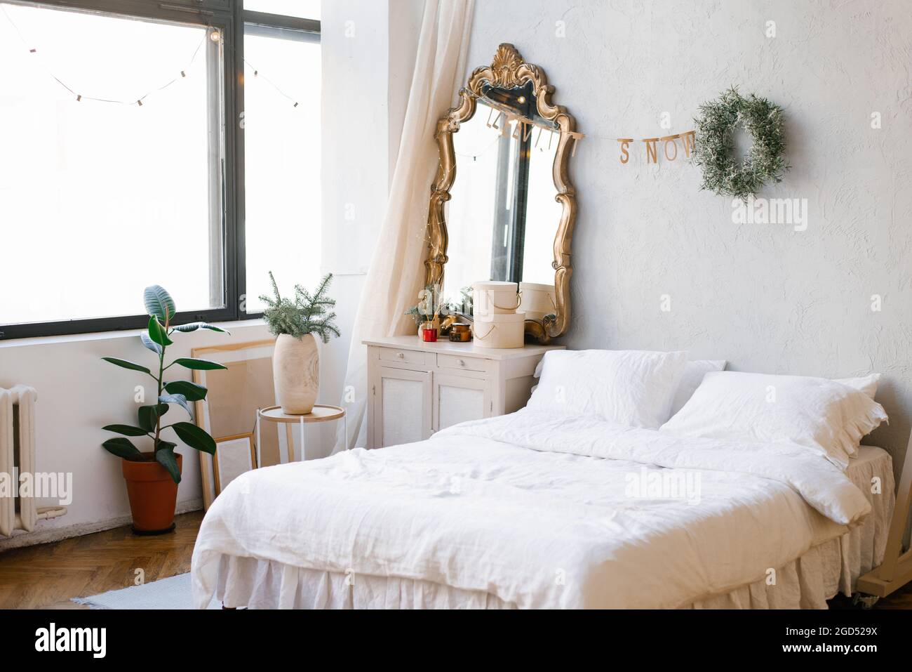 Un letto matrimoniale con lenzuola bianche e cuscini nella camera da letto decorata per Natale e Capodanno. Corona di Natale vicino allo specchio presso il comò Foto Stock