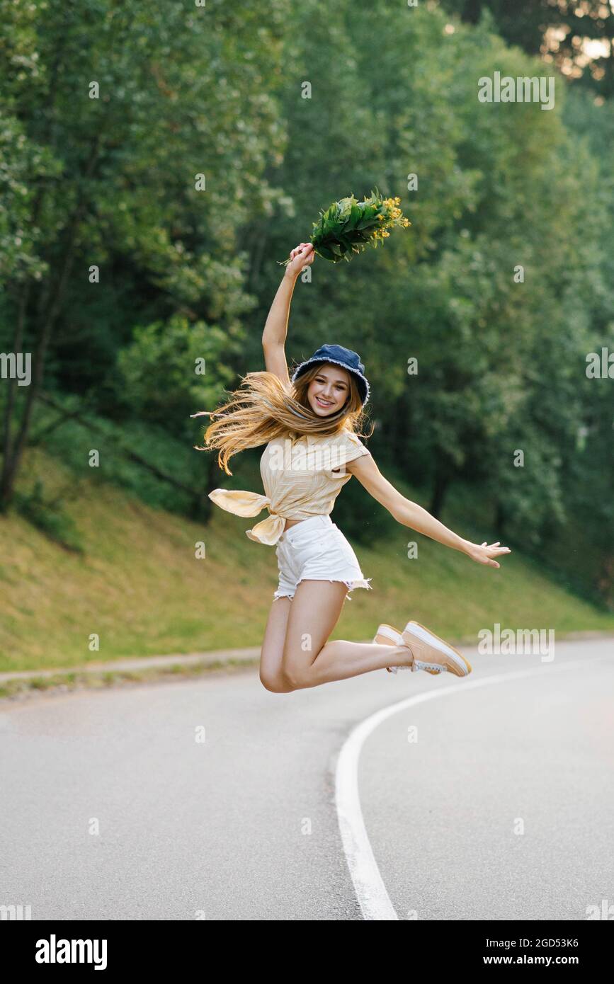 Una ragazza felice in pantaloncini bianchi e una blusa gialla salta con un bouquet di fiori, in piedi sulla strada vicino alla foresta Foto Stock