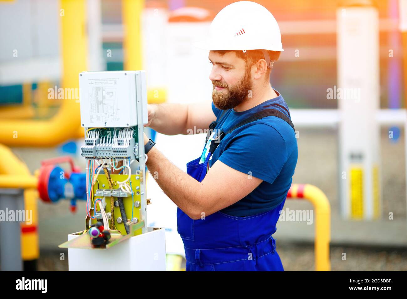 Un ingegnere elettrico qualificato crea un dispositivo di controllo e misurazione in un impianto industriale in una giornata estiva. Ingegnere specializzato in casco e uniforme sul lavoro. Un ritratto autentico. Foto Stock