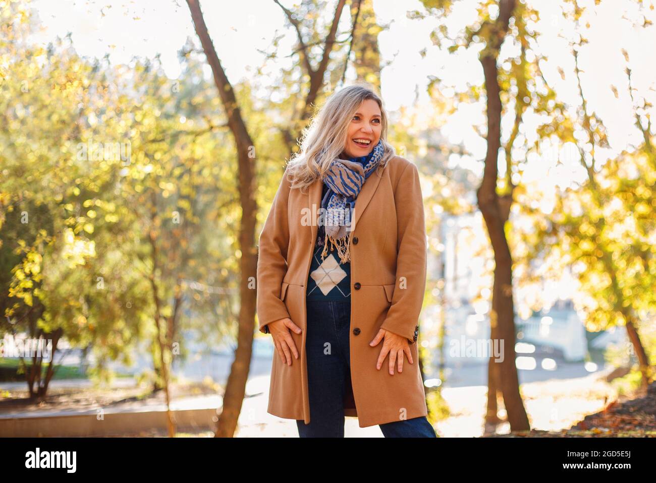 Felice donna di mezza età godendo l'autunno fuori, vestito con abiti casual e cappotto beige in piedi nel parco giallo, guardando da parte e sorridente allegro Foto Stock