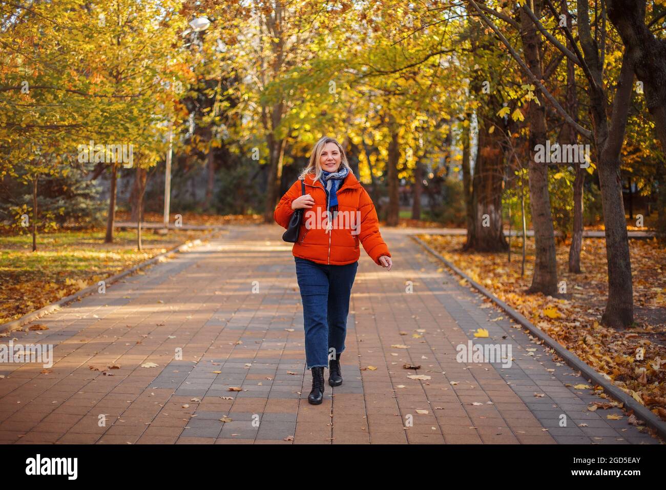 Felice positiva donna di mezza età in giacca arancione giù sorridente alla macchina fotografica mentre cammina all'aperto nella soleggiata giornata invernale, allegra donna matura godendo l Foto Stock